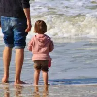A father and small girl stand on the sand, watching ocean waves with their feet in the water.