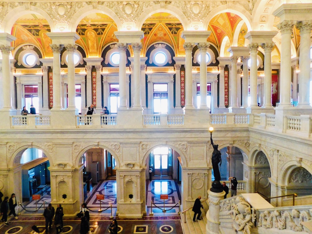 The atrium of the library congress as seen from a balcony with its marble stairs and columns, vaulted murals and light from windows.