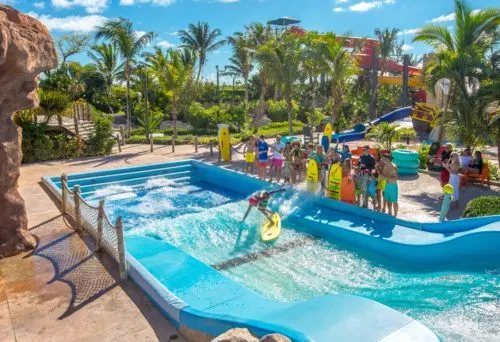 a group of kids watch another teen on the wave machine at beaches resort.