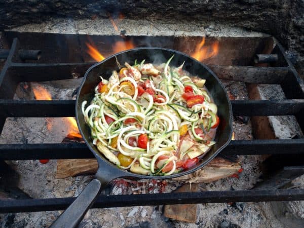 A cast iron skillet, full of chicken and veggies, cooking over a campfire.