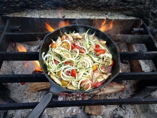 a cast iron skillet, full of chicken and veggies, cooking over a campfire.