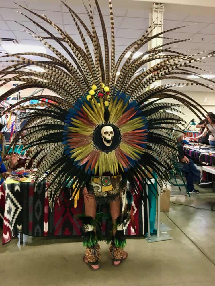 A man in an enormous featherd costume with a skull on the bank peruses the market at the gathering of nations.