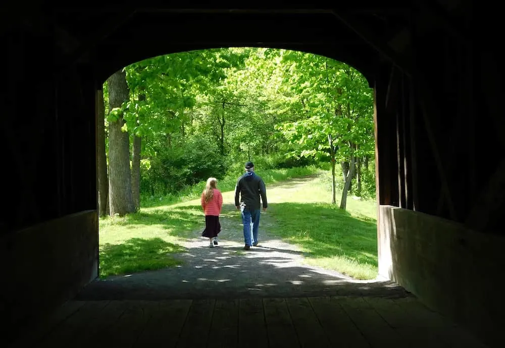 a girl and dad leaving the covered bridge on a nature trail in glimmerglass state park.
