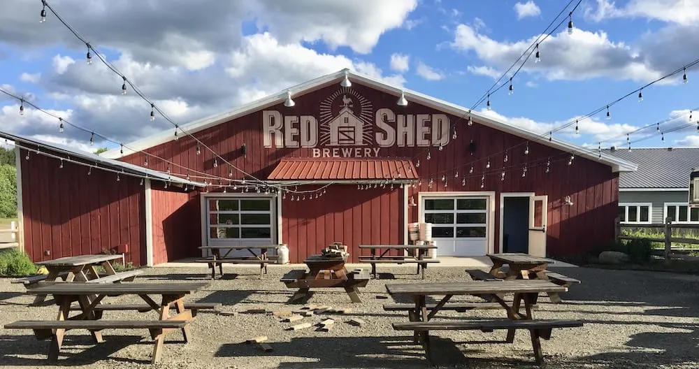 the barn-like red shew brewery with its beer garden in front it; it has picnic tables and lights.