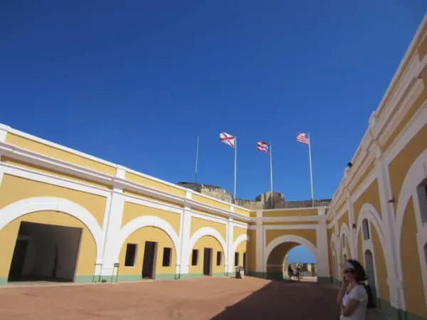 the colonial courtyard at the entrance to puerto rico's el morro fort has yellow plaster walls and both the puerto rican and american flags flying.