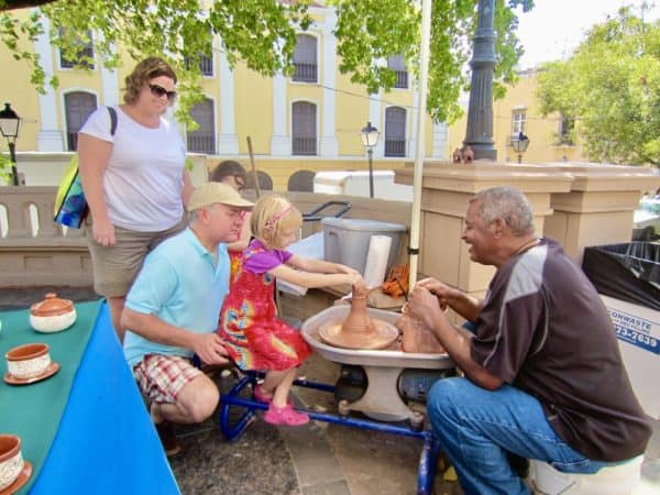 A young girl spins clay on a pottery wheel at a street stall in san juan, puerto rico while the owner and passersby watch.