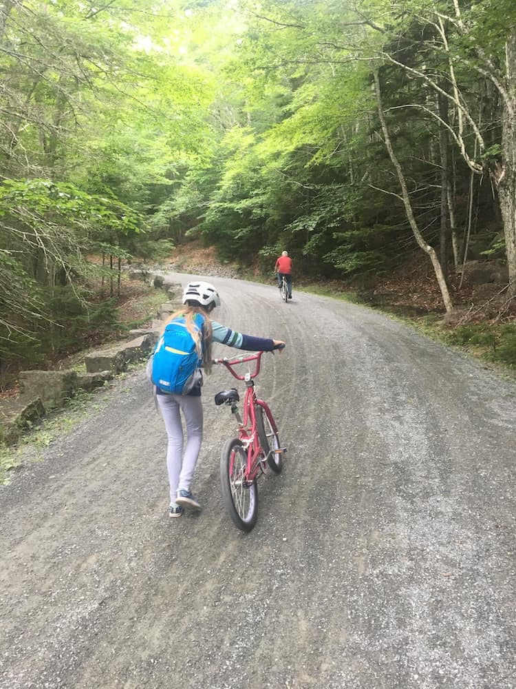 a girl walks her bicyle up one of the many long hills on the carriage roards in woodsy acadia national park.