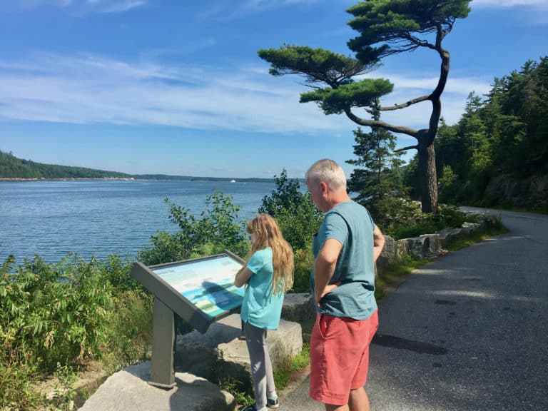 A girl and her dad read a sign on the wooded shore of acadia, looking for their first earthcache clue.