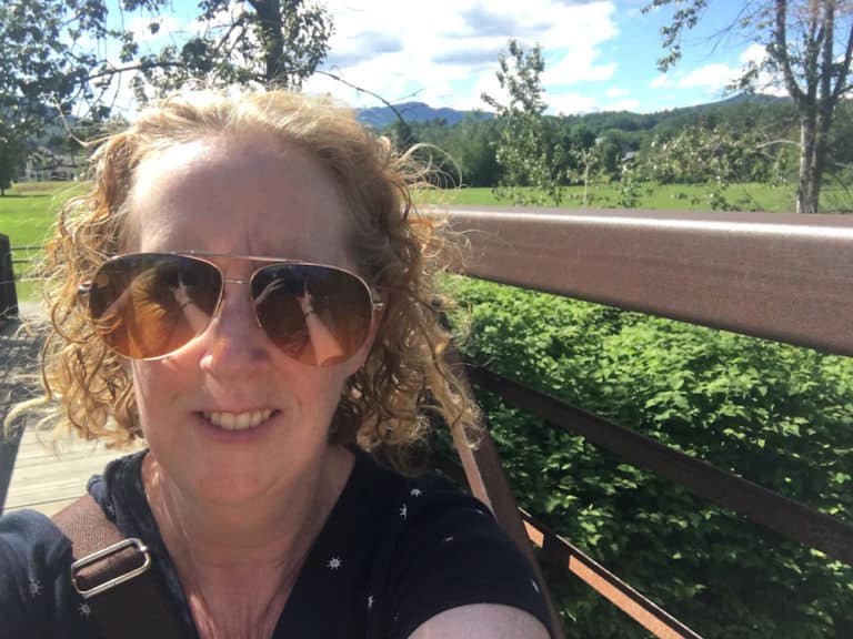 A woman takes a selfie from a bridge on the stowe recreation trail with fields and the green mountains behind her.