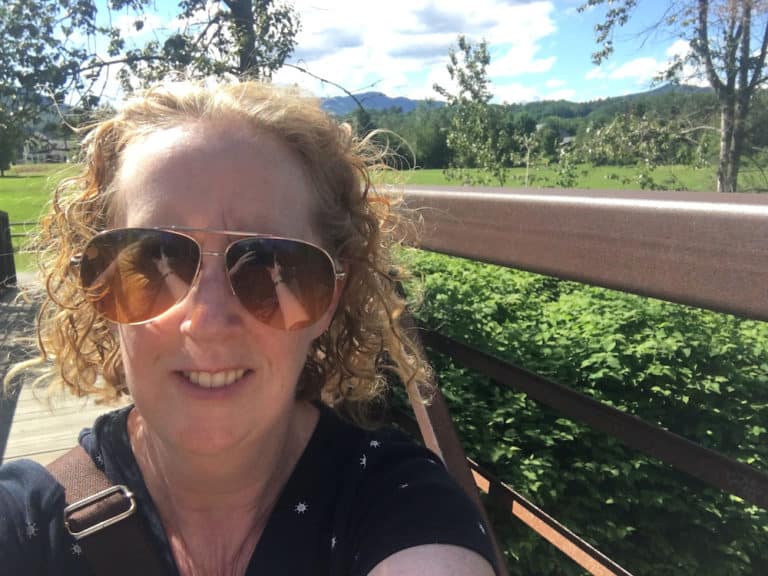 a woman takes a selfie from a bridge on the stowe recreation trail with fields and the green mountains behind her.