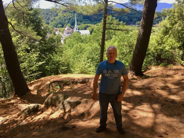 A man poses on sunset rock with trees and downtown stowe's whitel church steeple in the background.