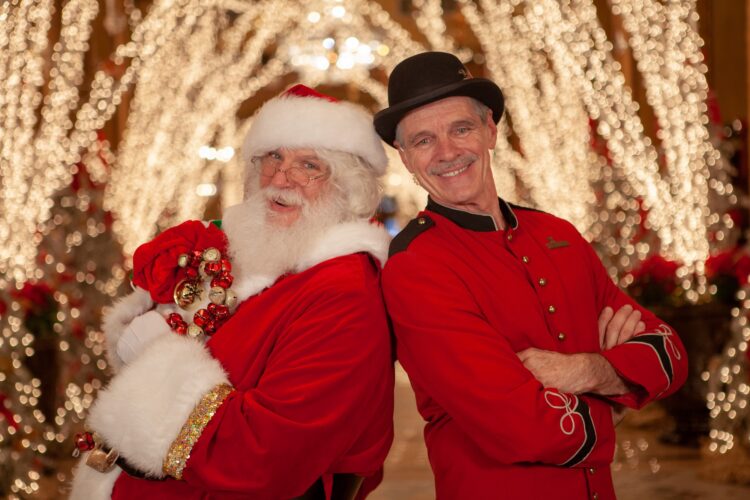 Santa claus and a doorman in red uniform and bowler hat post with birch brances and white holiday lights behind them.