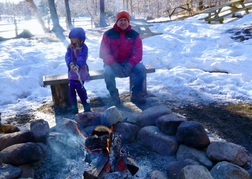 a girl avoids the smoke while toasting marshmallows over the fire at high falls gorge while her dad looks on. 