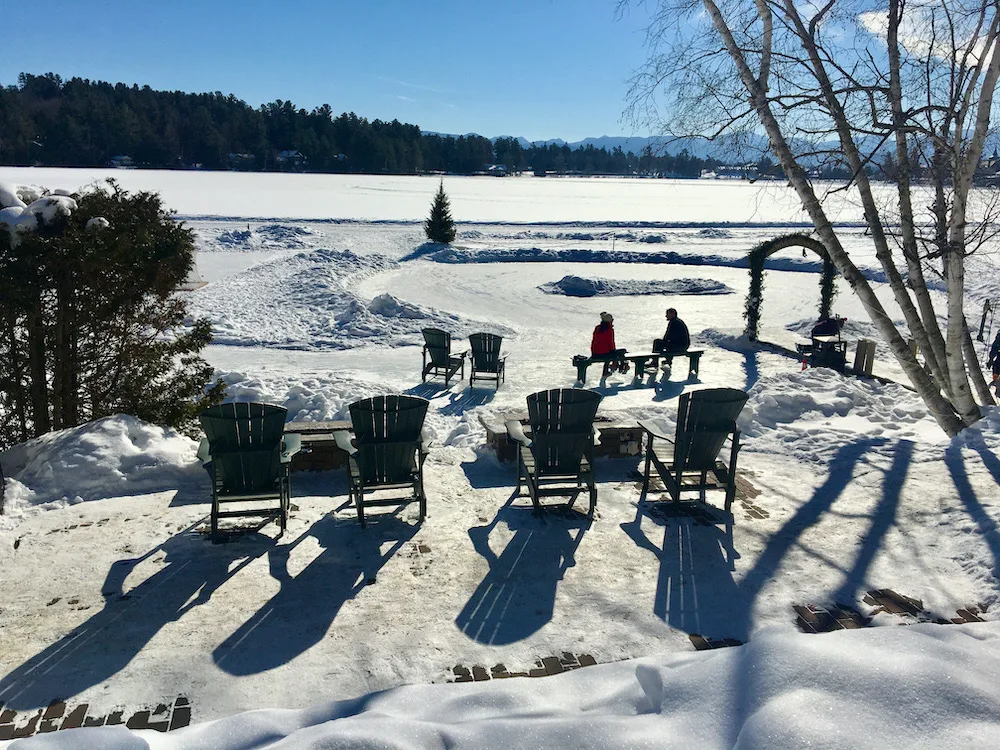 a couple sits on a bench on next to a cleared skating rink on from mirror lake in lake placid