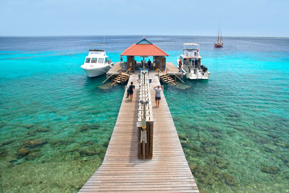 A dad and teenager walk down a pier in the blue caribbean sea toward two fishing boats. Divi resorts are good choice for single parents looking to vacation affordably with their kids.