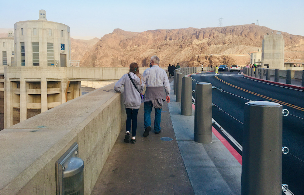 A dad and daughter walk across the top of the hoover dam with mountains in the background