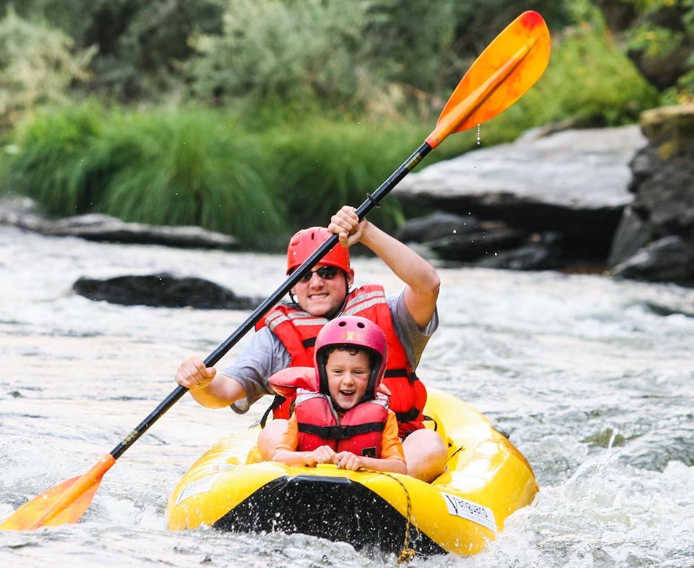A dad and small son in helmets and lifevests have fun paddling a rugged inflatable kayak down river rapids l a oars vacation