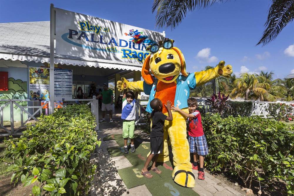 An interracial group of kids hugs raggs, yellow dog that is the mascot for divi resorts, a family friendly brand that is priced fairly for single parents.