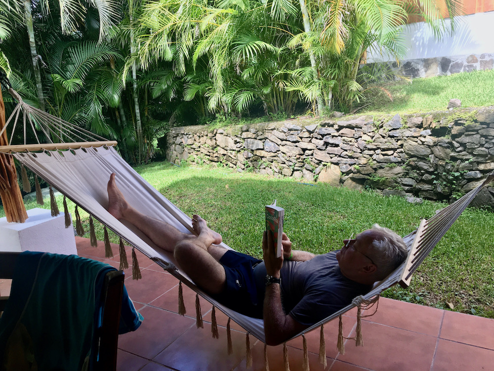 a guest at hacienda guachipelin reads a costa rica guidebook in a hammock on a flagstone porch surrounded by a garden.