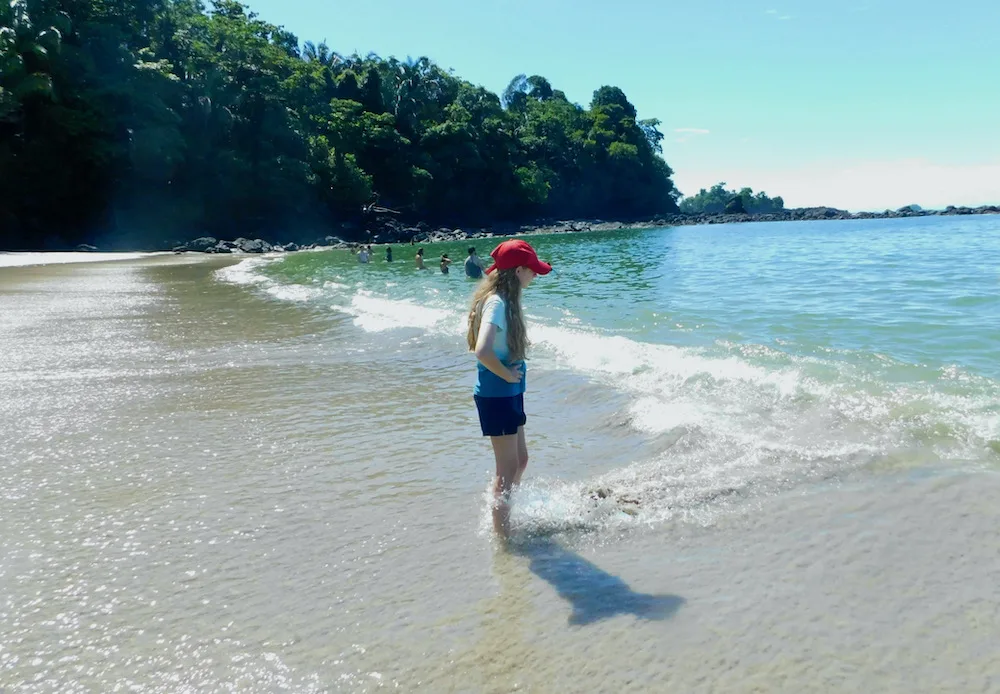 a girl stands with her feet in pacific ocean waves at cove beach in manuel antonio national park