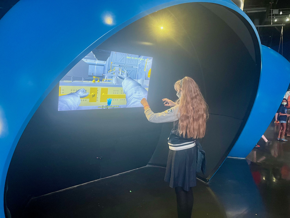 At the space shuttle building at cape kennedy visitor's center a teen tries her hand at making remote repairs with robotic arms that mimic here movements on a screen.