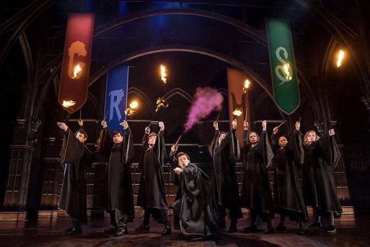 A row of hogwarts students in black robes get their wands ready to duel -- purple smoke comes from the center character -- in harry potter & the cursed child