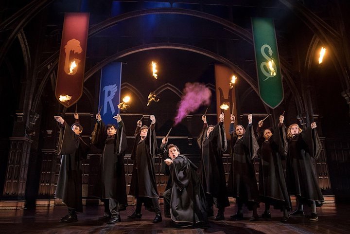 a row of hogwarts students in black robes get their wands ready to duel -- purple smoke comes from the center character -- in harry potter & the cursed child