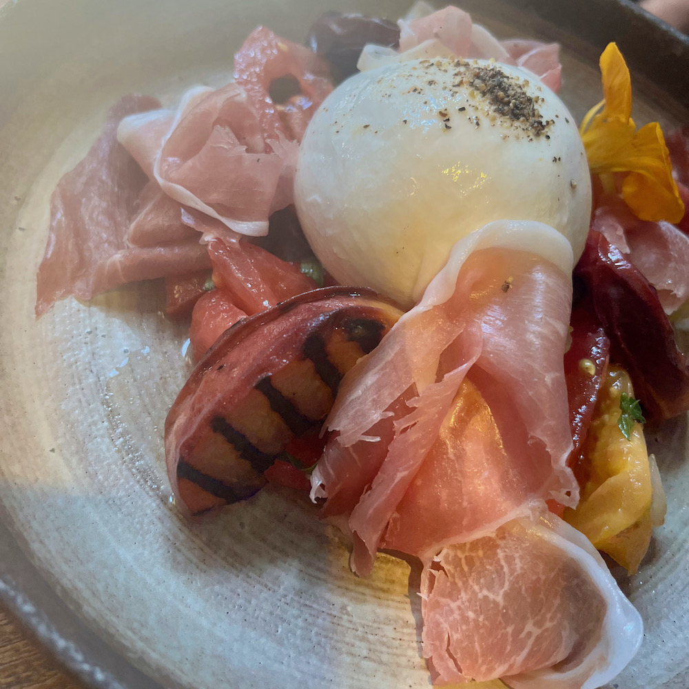 at the caxtion grill, a summer salad has burrata surrounded by proscituuo, tomato and grilled peaches.