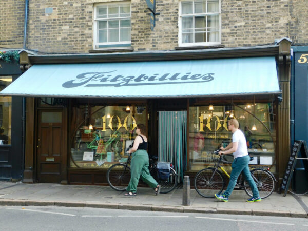 Fitzbillies café is an essential stop on any visit to cambridge with teens. Two people with bikes pass under its blue awning.
