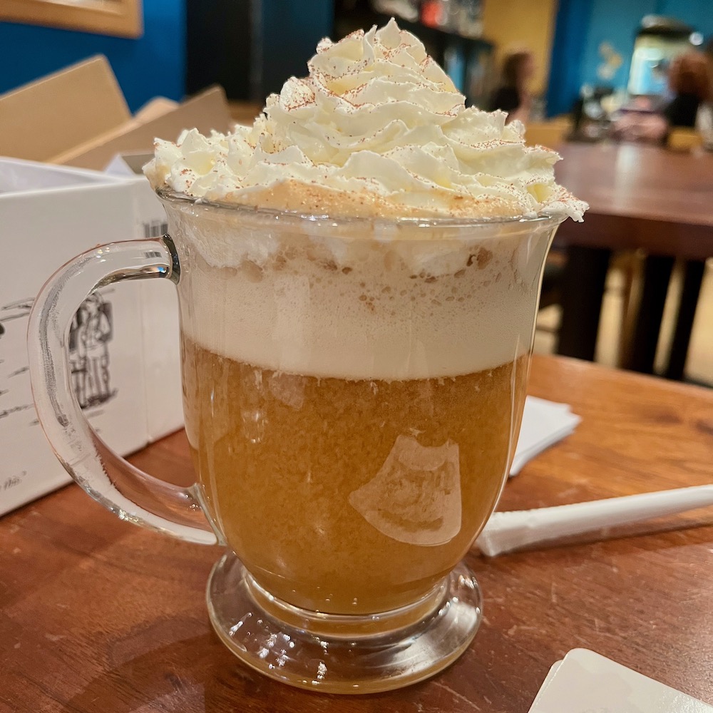 a glass mug of warm butterbeer with a head of whipped cream on top with a game café in the background.
