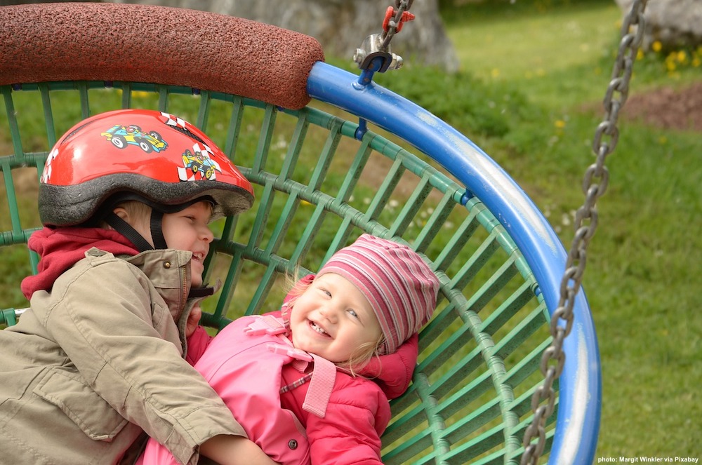 Two toddlers giggle on a large nest swing. Playgrounds are a great resource when you take an active toddler on a vacation