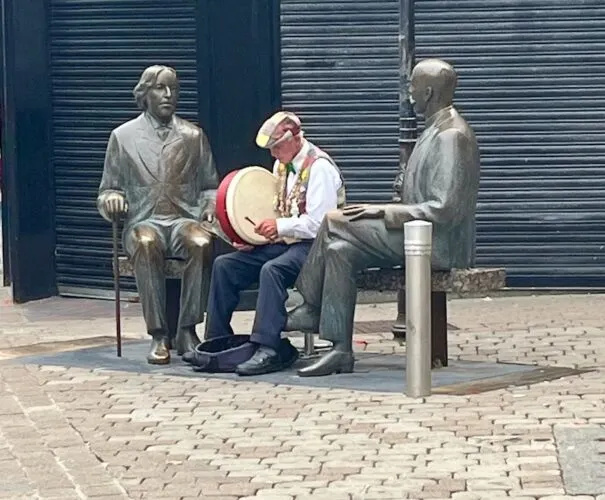 a man in a cap and waistcoat plan tthe bodhran next to a statue of oscar wilde in galway.