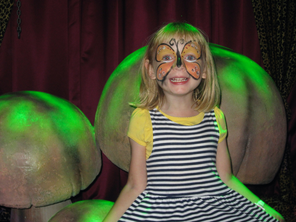 a girl with a butterfly mask painted on her face sits on giant fake mushrooms on the norwegian breakaway