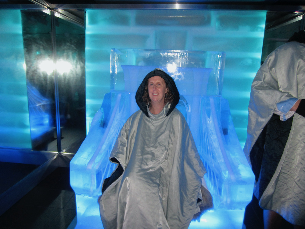 a woman sits on a large chair made of ice with a hooded thermal cloak in the ice bar on the ncl breakaway.