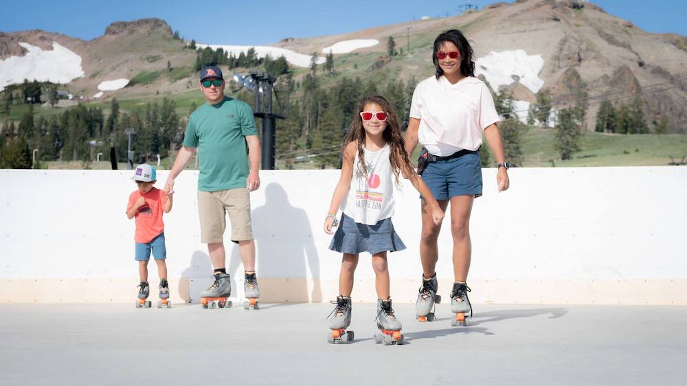 A family roller skates with snow in the mountains behind them during the summer at the high camp in lake tahoe.