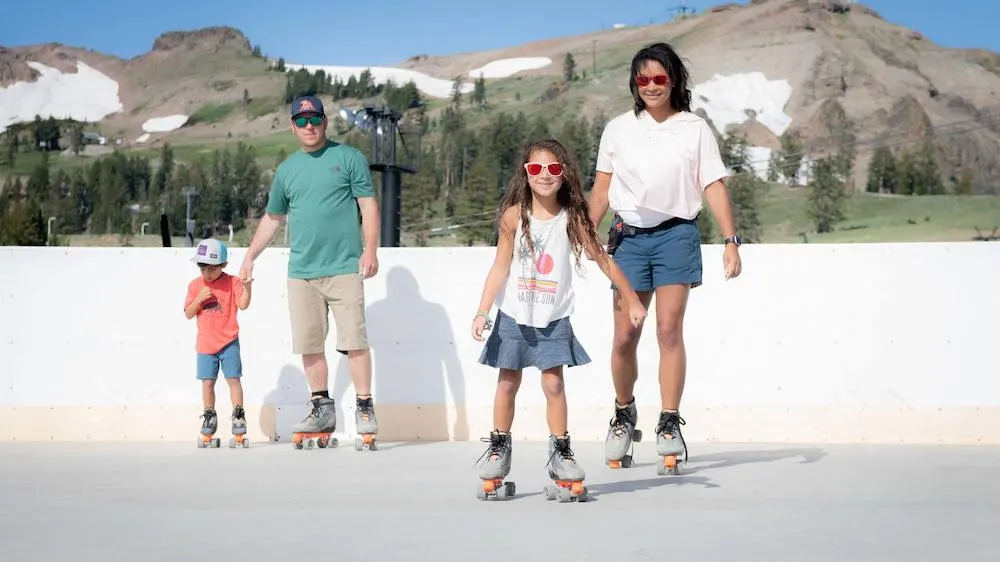 a family roller skates with snow in the mountains behind them during the summer at the high camp in lake tahoe.