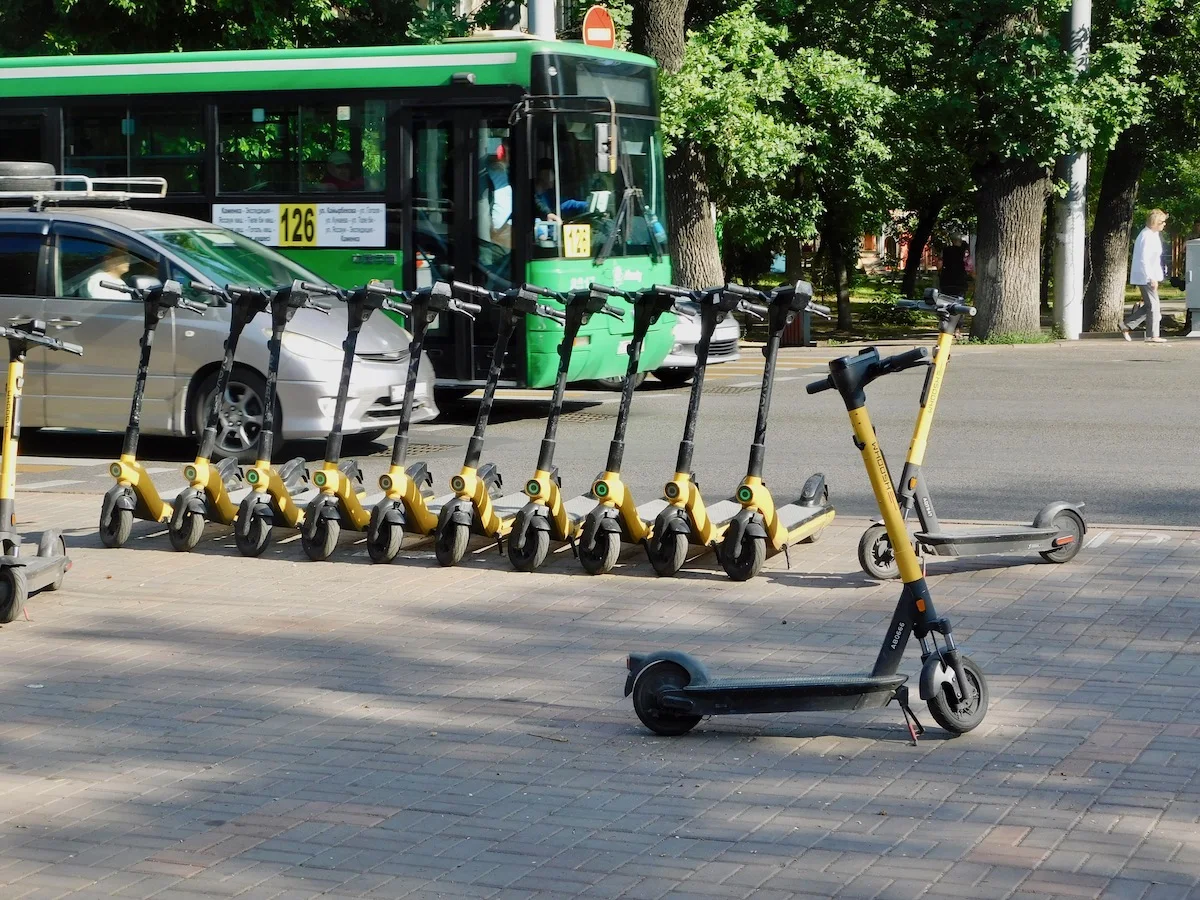 Travel To Kazakhstan With Kids? Here's All You Need To Know: Scooters wait on an Almaty sidewalk to be rented as buses and cars pass on a busy street.