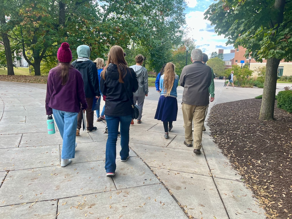 A group of high schoolers and their parents follow a university of rochester senior in a blue school jacket on a tour of the campus.