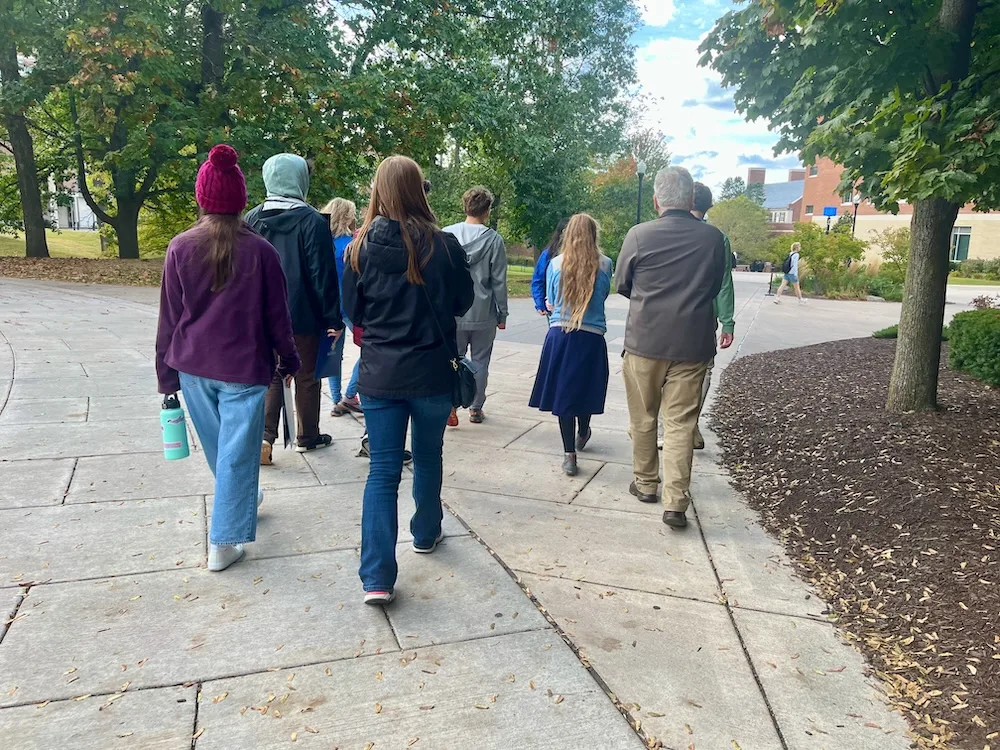 a group of high schoolers and their parents follow a university of rochester senior in a blue school jacket on a tour of the campus.