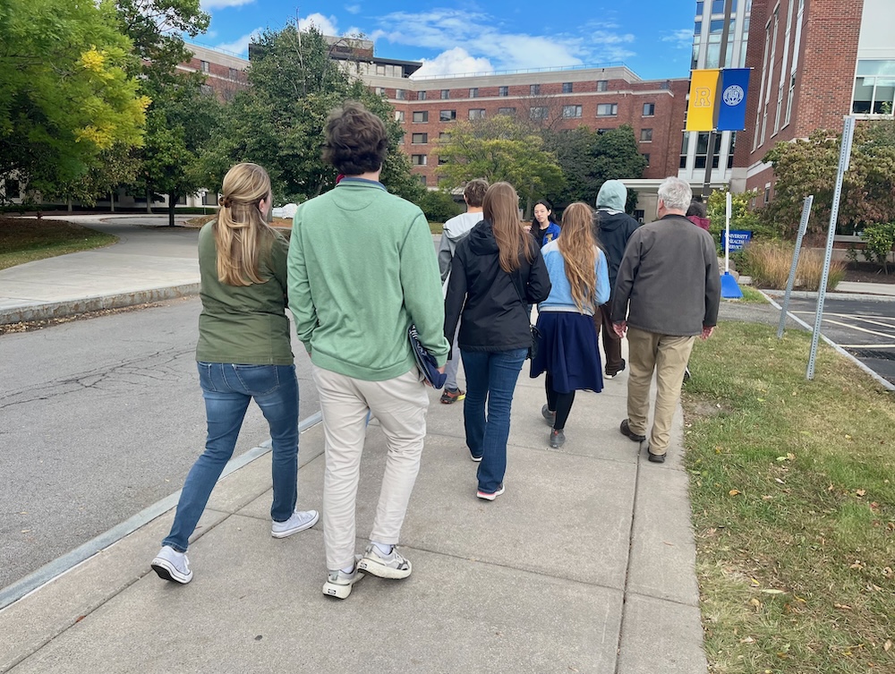 a group passes between a quad and a parking lot on a university of rochester high school admissions tour. 
