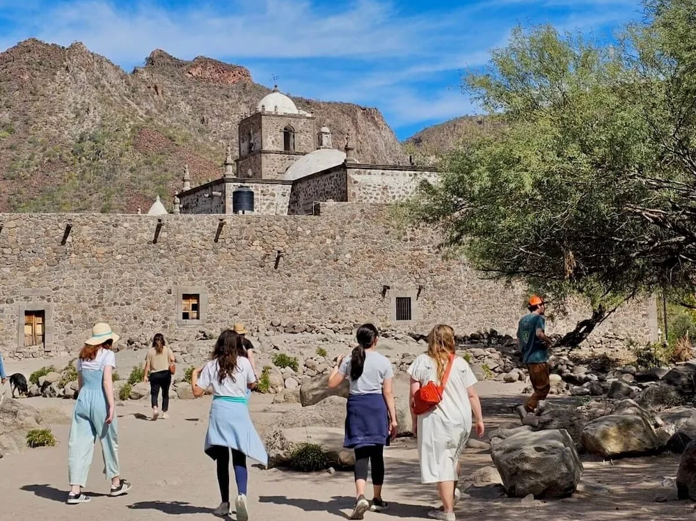 a group of tens walk past rocks and cypress trees toward mission javier in the foothills above loreto.