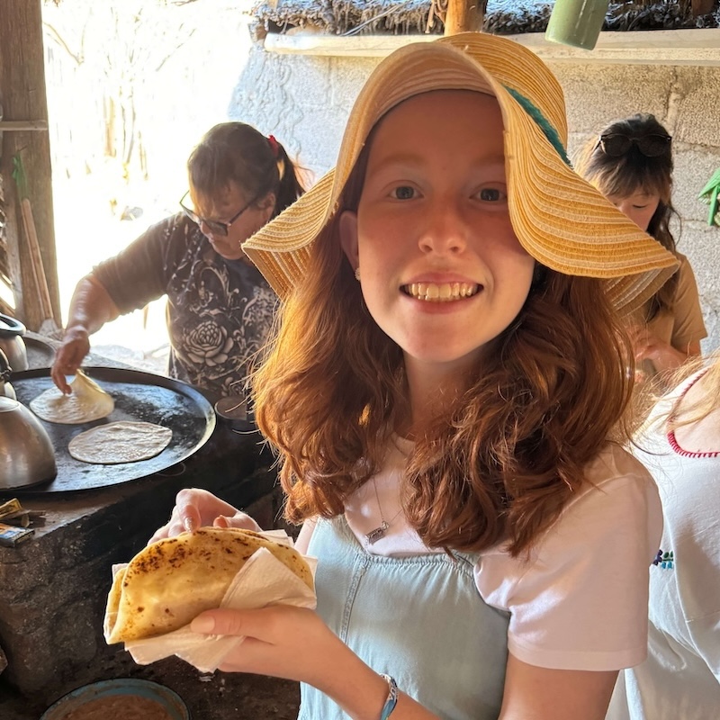 A girl eats a warm homemade tortilla while a ranch owner cooks more on a griddle.