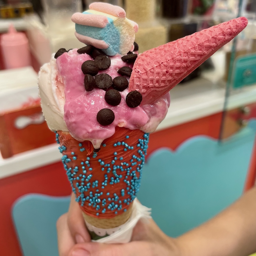 A colorful and elaborate ice cream cone from hans & gretel in monastiraki has pink ice cream, cookie bits, a pastel marshmello and a tiny pink cone hat.