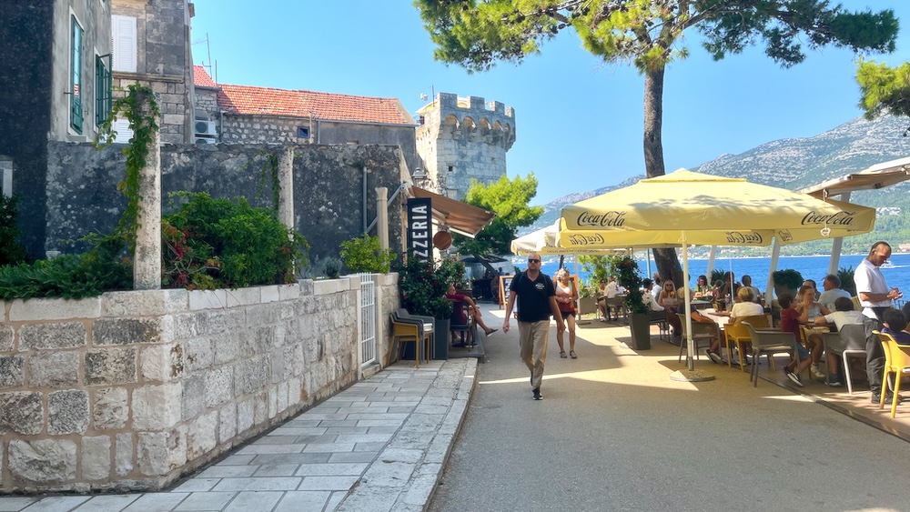 the outdoor restaurant tables in korcula's old town have medieval stone buildings behind them, the blue adriatic sea below them and mountains in the distance. 
