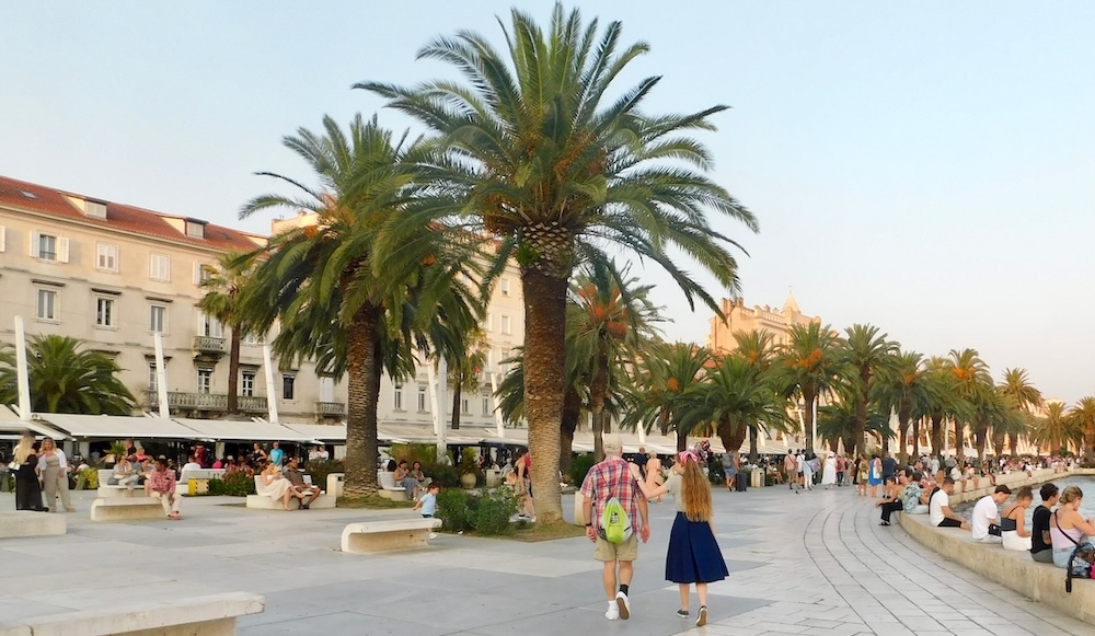 a dad and teen girl stroll on split's water-front promenade with palm trees, shaded outdoor cafés and historic red-roofed buildings behind it. it's a mediterranean cruise port teens like.