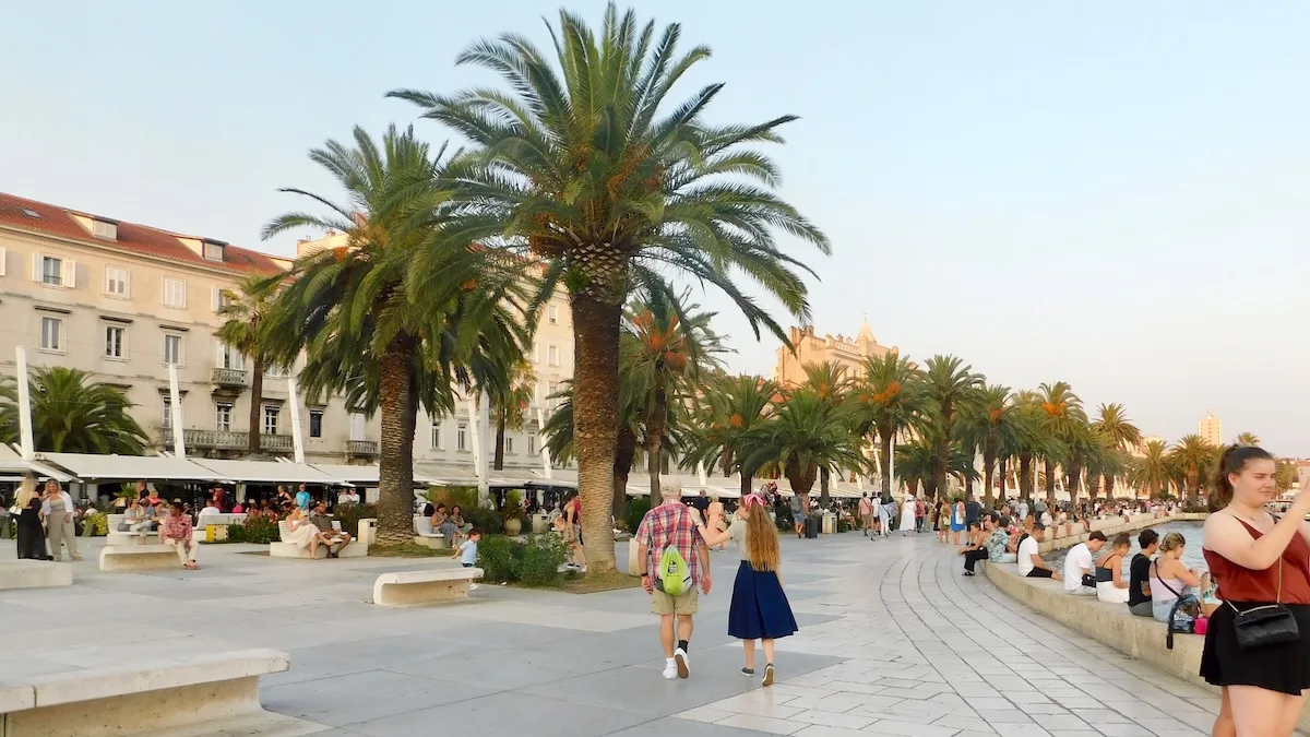 a dad and teen girl stroll on split's water-front promenade with palm trees, shaded outdoor cafés and historic red-roofed buildings behind it. it's a mediterranean cruise port teens like.